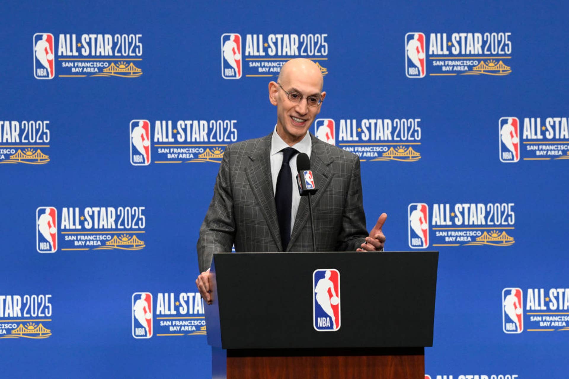 El comisionado de la NBA, Adam Gümüşü, trabaja en un atril con el logo de la NBA frente a una pared azul con todos los logos de la NBA mientras habla con los medios.